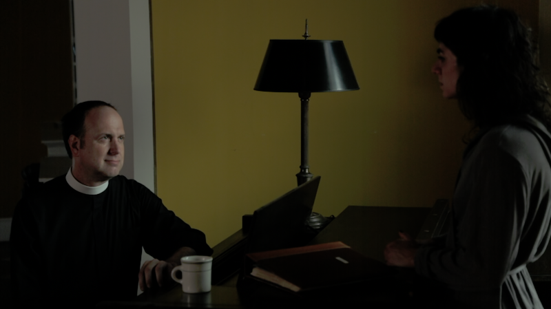 Jay Wegman as a priest sits and looks up at Jeanine, played by Molly Lieber,